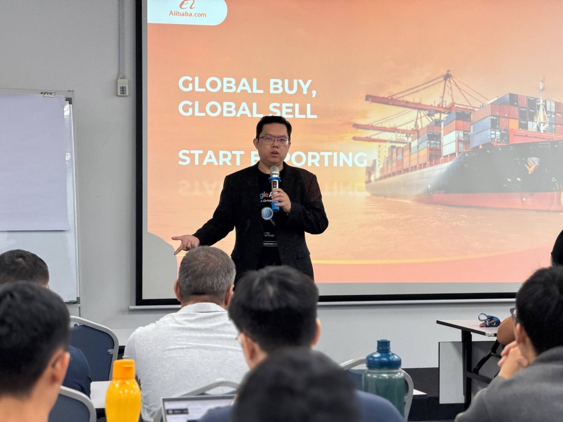 A speaker presenting to a seated business audience in front of a screen reading Global Buy, Global Sell and Start Exporting