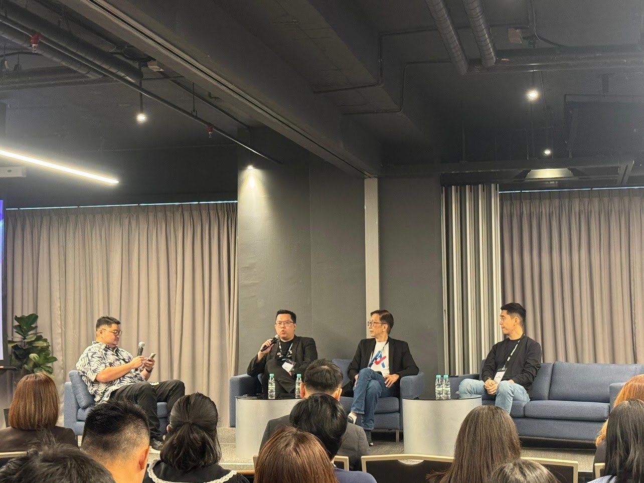 A panel discussion with four seated speakers on a stage in front of an audience, in a dark room with curtained walls