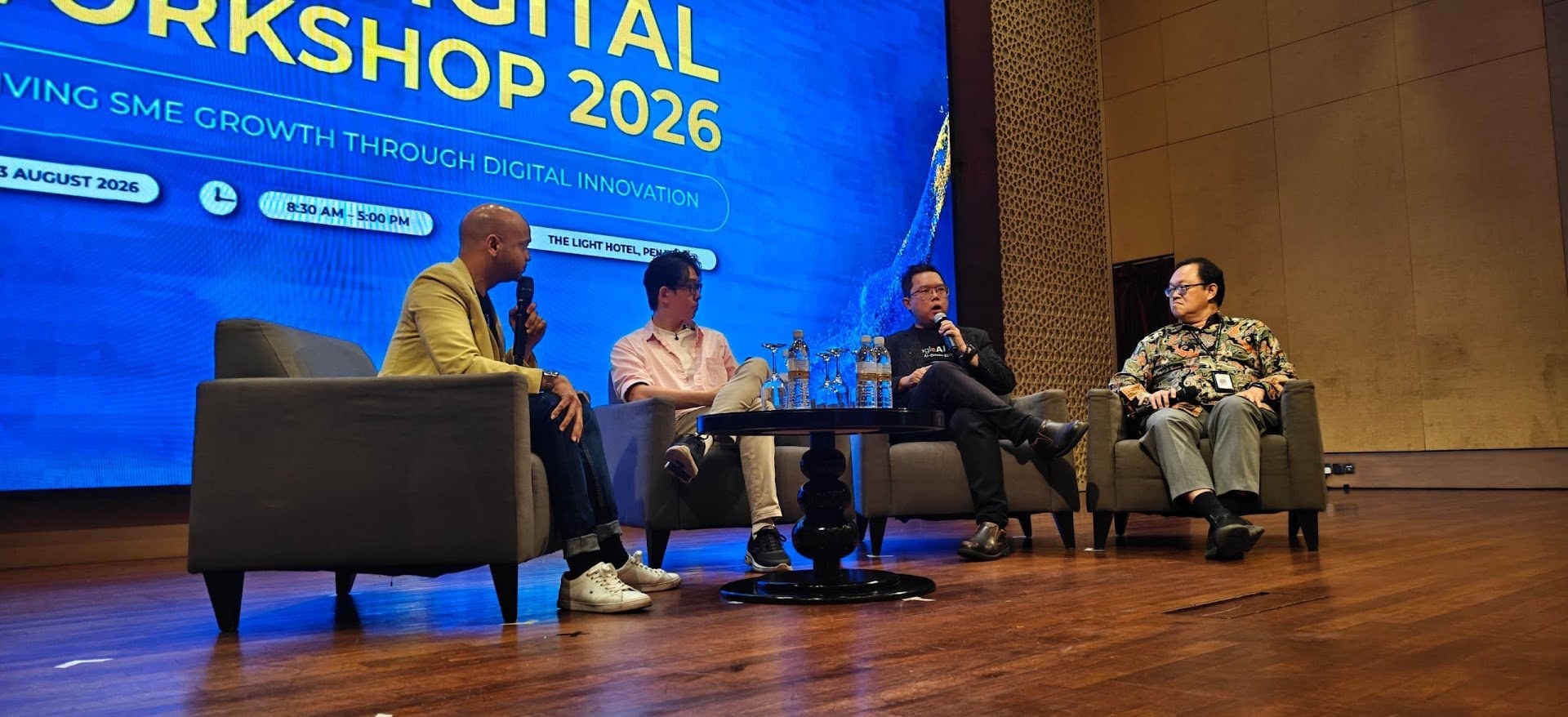 A four-person panel on stage at a digital workshop in 2026, seated in armchairs beneath a large blue event screen reading Digital Workshop 2026