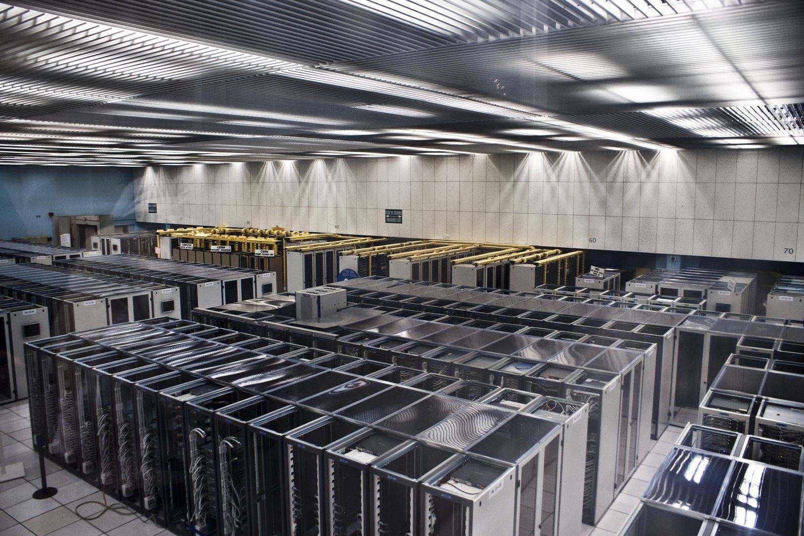 A large data centre hall filled with rows of server racks and overhead cable trays, lit from above