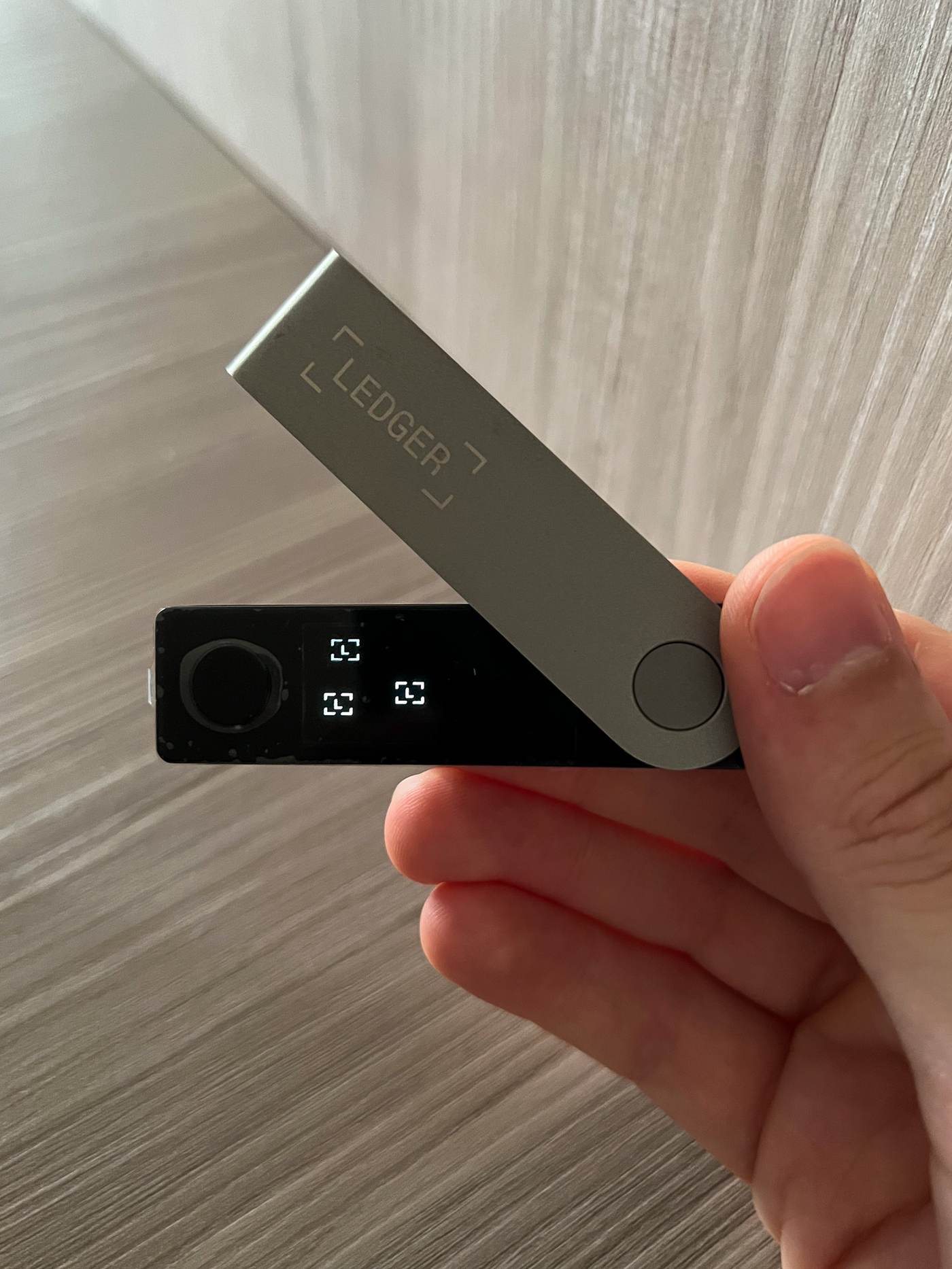 A hand holding an unmodified Ledger Nano X hardware wallet, showing the screen and the device's own casing